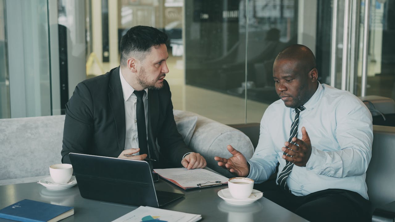 heros-img Two businessmen in discussion over coffee and laptop in modern office setting.